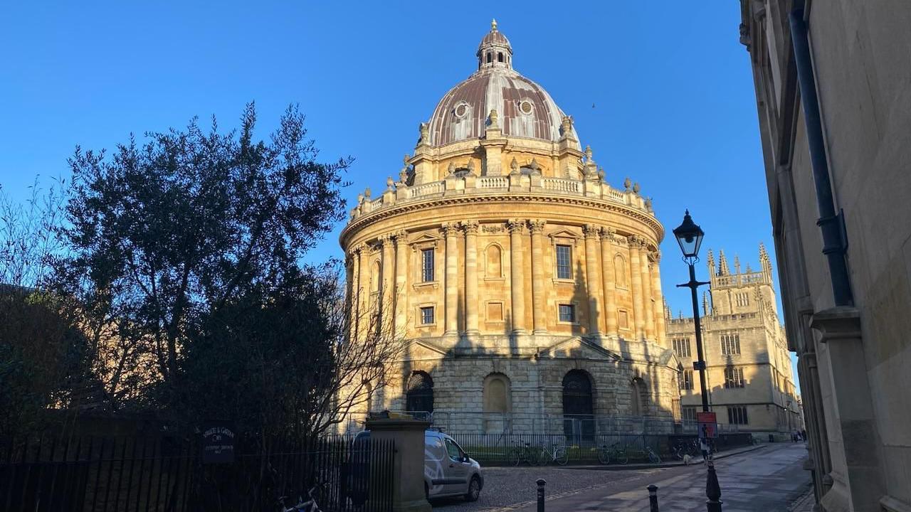 The yellow-stone building of the Radcliffe Camera is illuminated in the sun. The building is round with a domed roof. In the foreground is a tree in silhouette and a white van. 