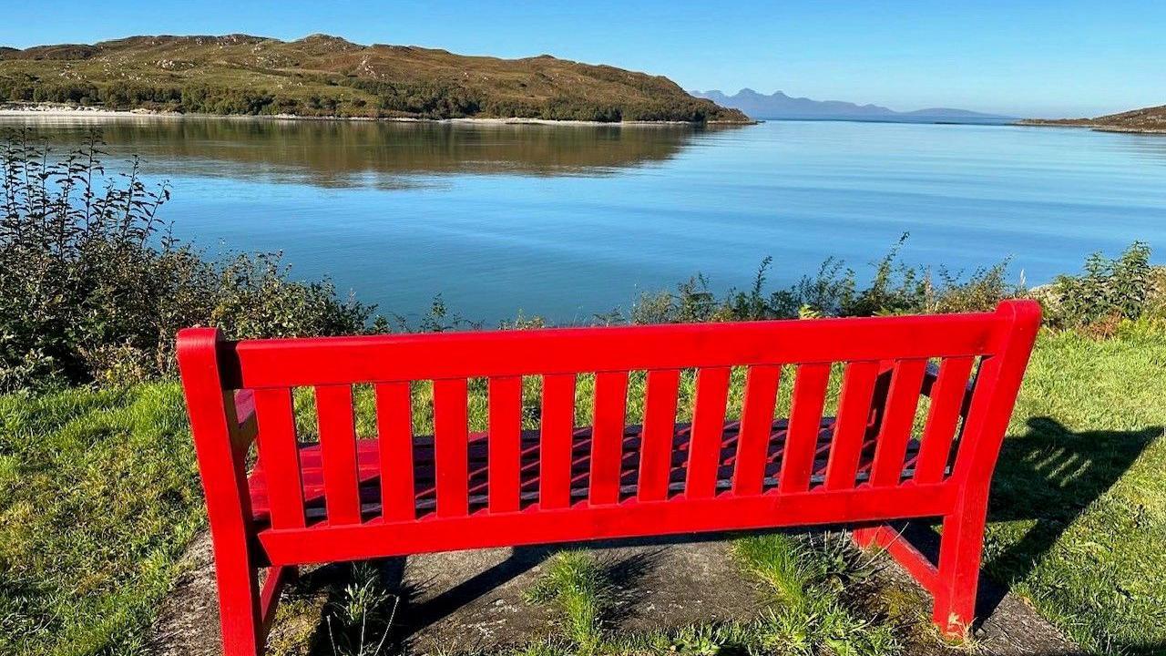 A red bench at the side of a body of water looking out at the reflection of a nearby land mass