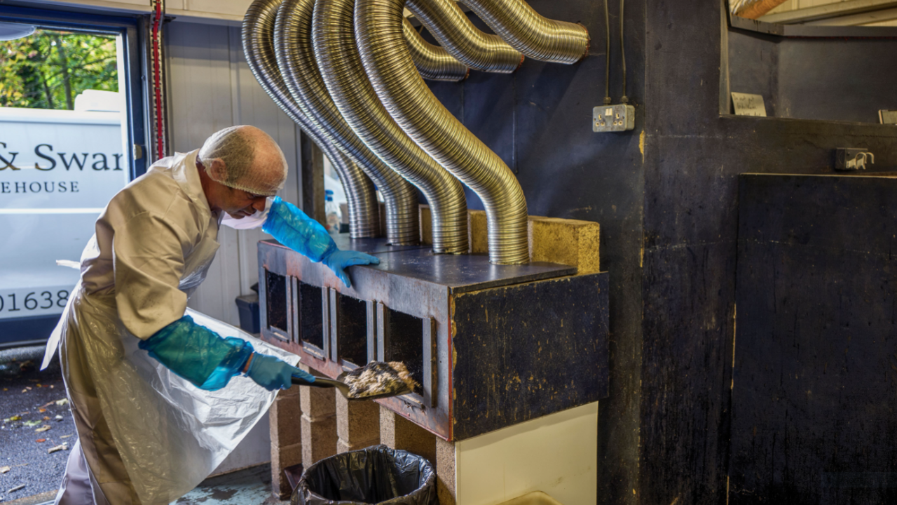 A man wearing a hairnet and white overalls and blue gloves putting what looks like salmon into a large oven