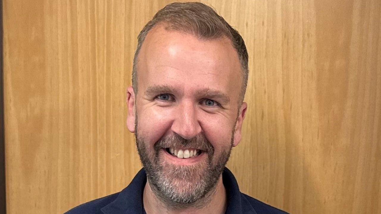 Headshot of Sam Brunt smiling at the camera, wearing a blue polo shirt.