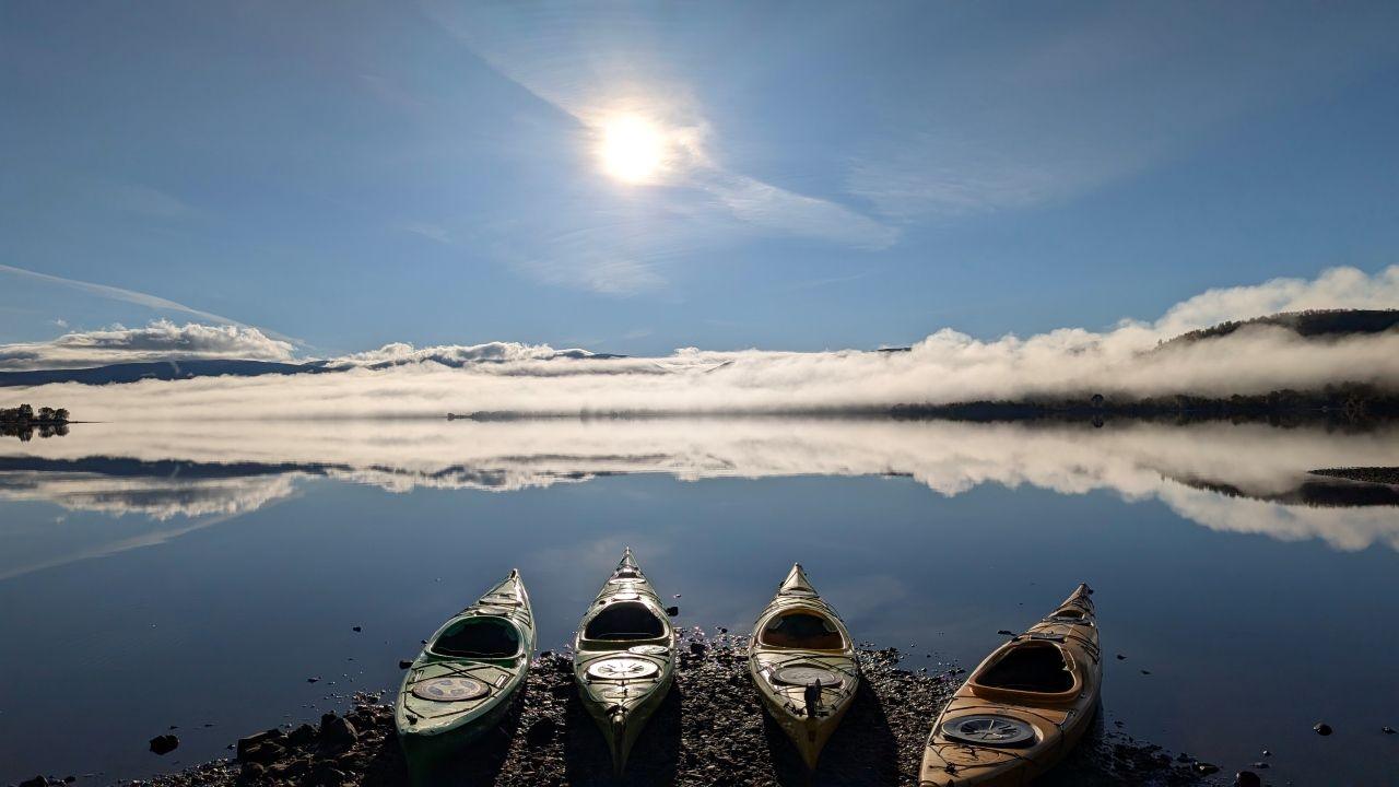 Four kayaks sit at the edge of a loch. Clouds are reflected in the water and there is a hazy sun above