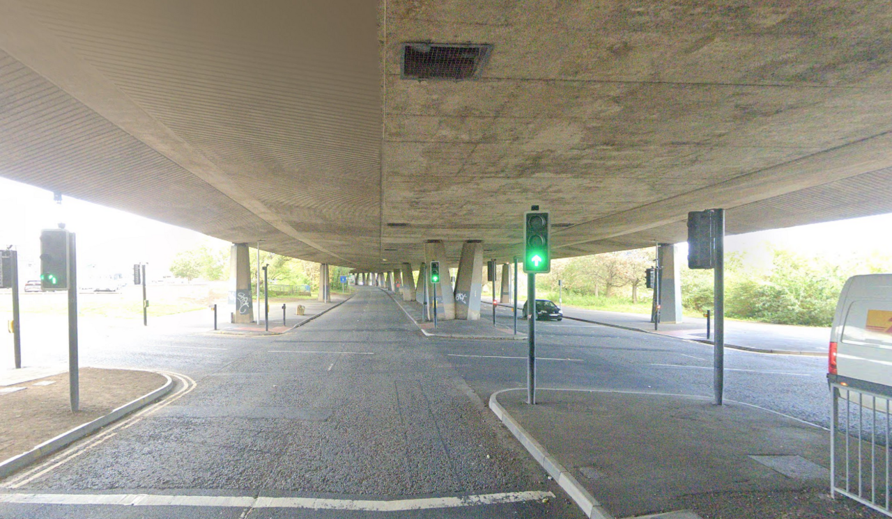 A huge concrete flyover held up by many pillars sits above two roads. There are traffic lights on green and a car is approaching them.