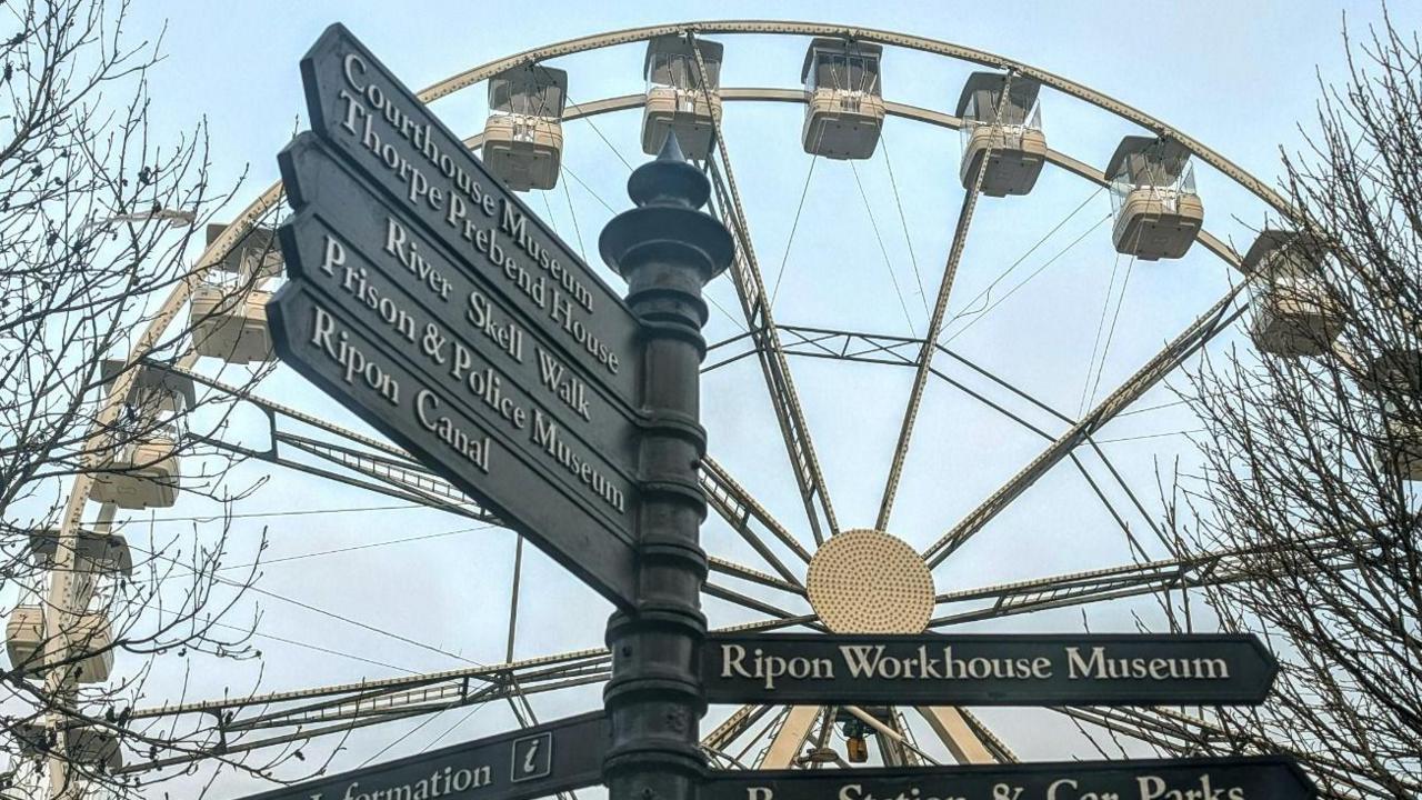 A green sign post with seven pointers displaying information about Ripon attractions with the big wheel behind it.