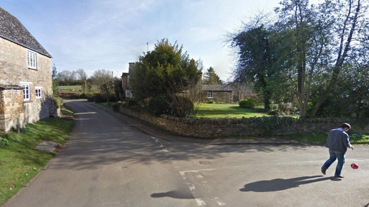 A screenshot from Google street view of a junction of two country lanes. A man is walking in the middle of one of the roads and there's a cottage at the junction with a large garden.