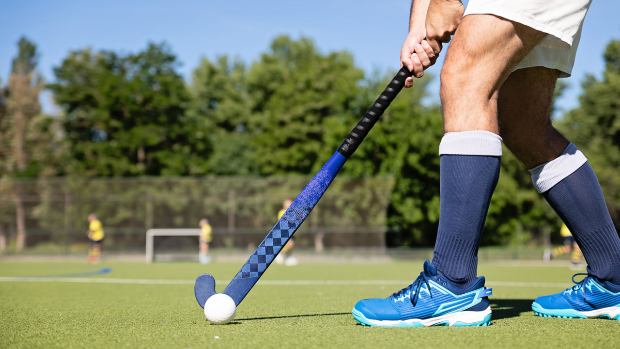 A hockey player holding a hockey stick next to a hockey ball. They are pictured only from the waist down. They are wearing white shorts, navy shocks up to their knees, and blue trainers. They are standing on a hockey pitch.