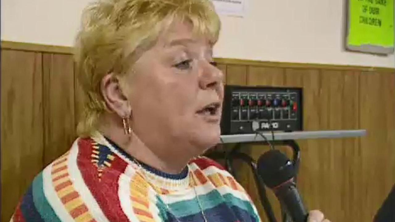 A screen grab of Haney addressing a public meeting. She is holding a microphone. She has short blonde hair and a big gold hooped earring. She is wearing a multi-coloured jumper