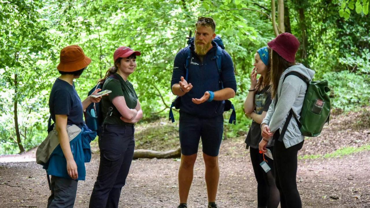 A group of walkers standing together in a clearing in woodland. Some are wearing backpacks, hats and waterproof jackets tied about their waist