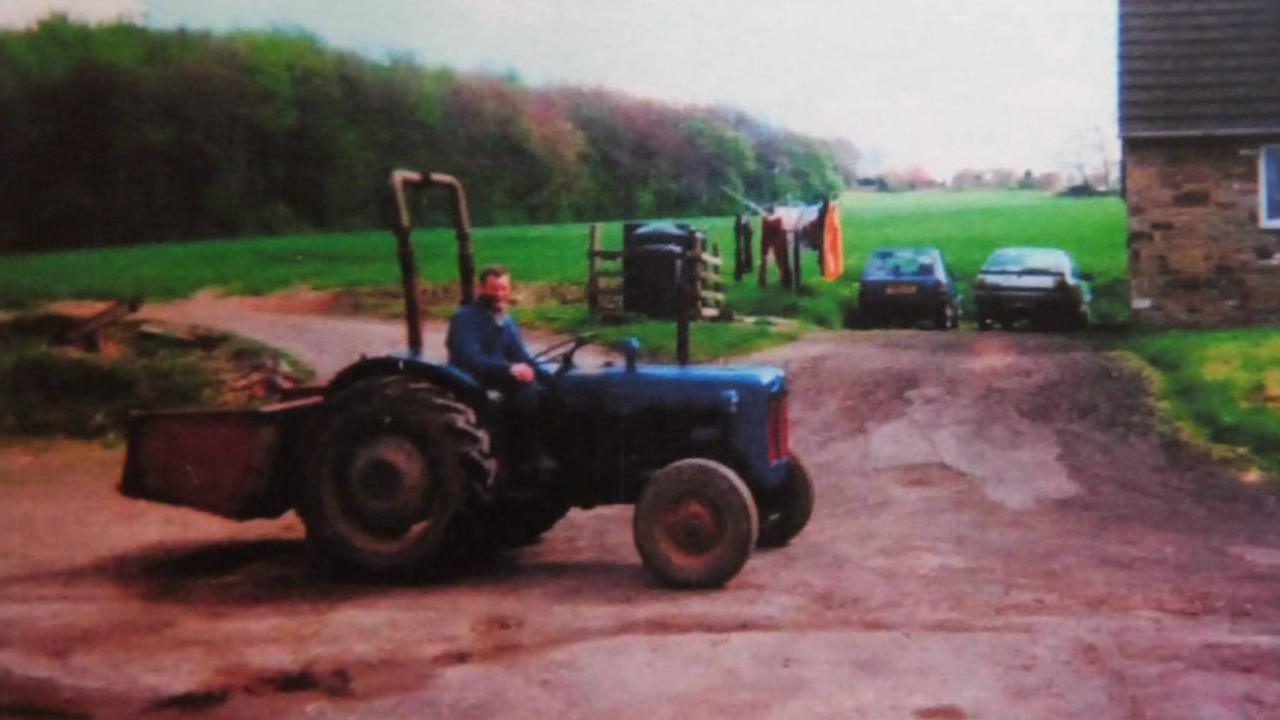 A man sits on a blue tractor, looking towards the camera. There is a house in the background, alongside two cars and a clothing line.