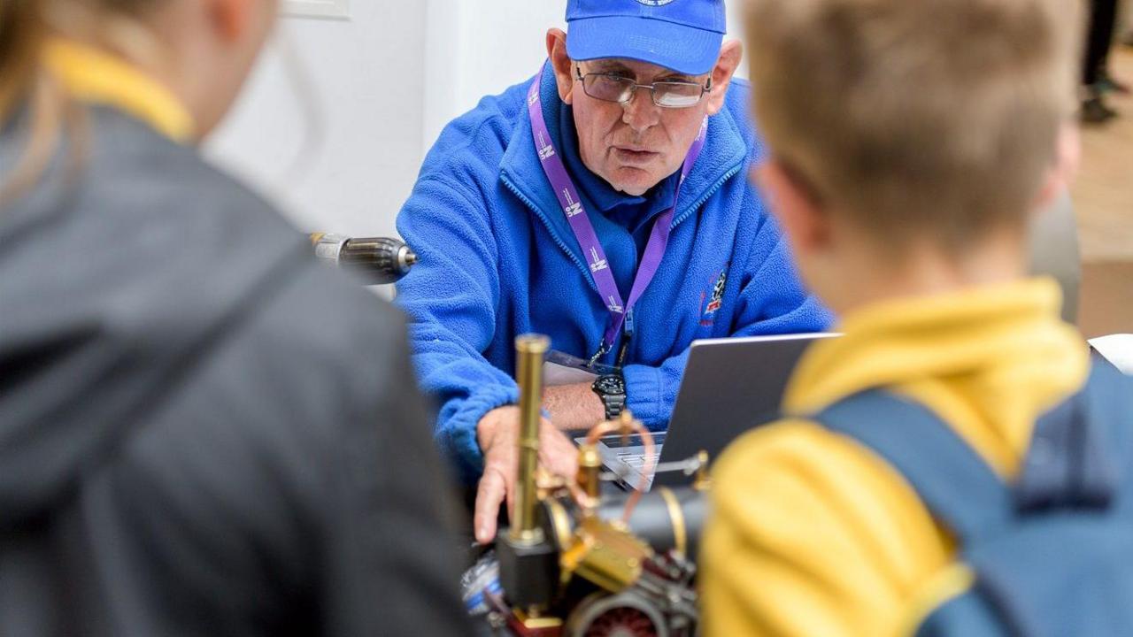 Man in blue overalls shows two pupils a steam engine