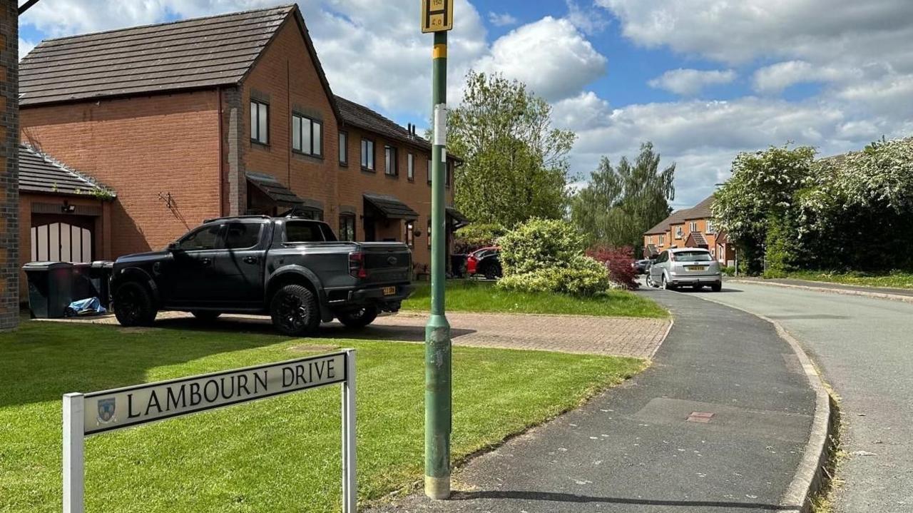A road with red brick houses on one side and cars parked on drives. Front gardens can be seen of green grass with the bottom of a green lamppost near the camera and in front of that a road sign saying "Lambourn Drive".