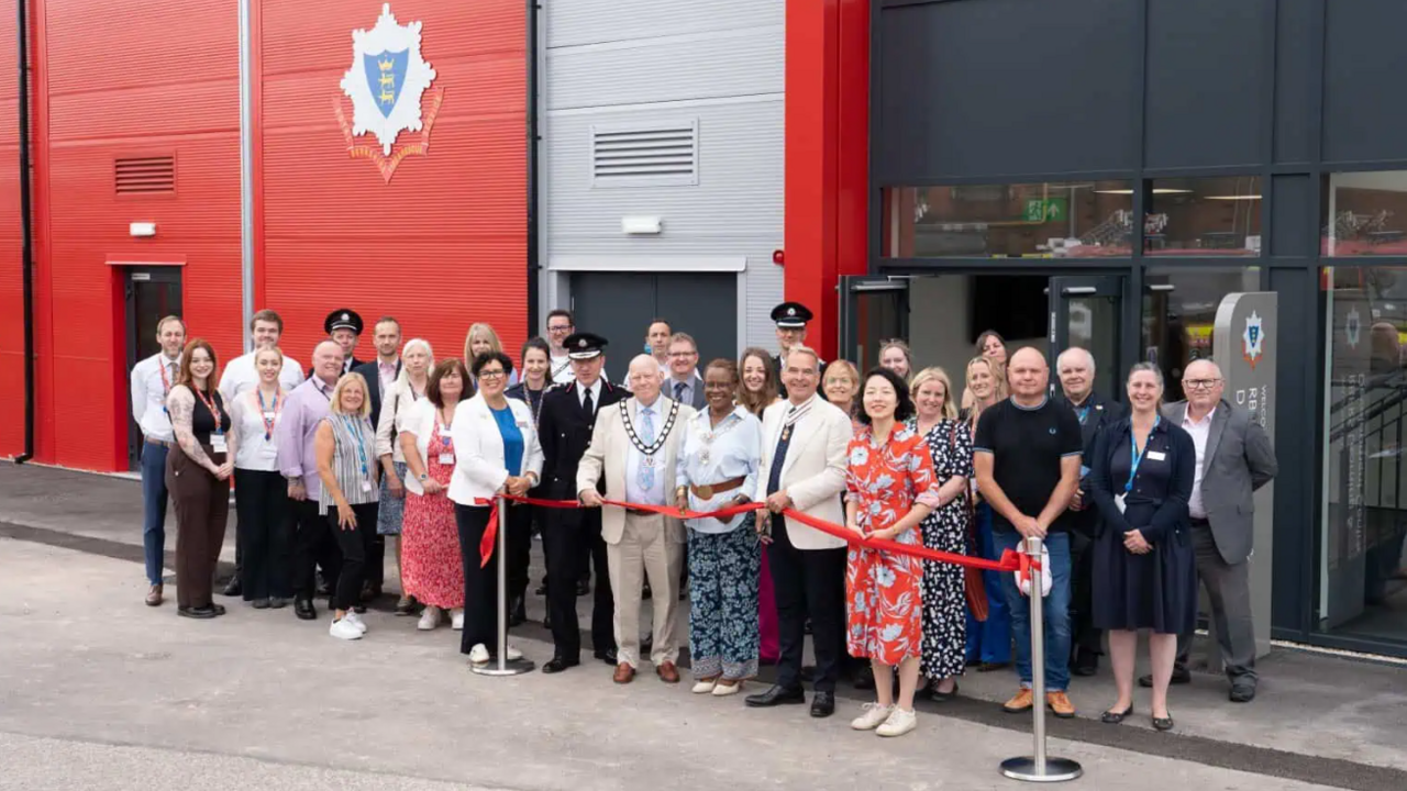 A large group of people, dressed smarty, standing outside the entrance of the new training centre. The building has red, silver and black walls and glass doors.