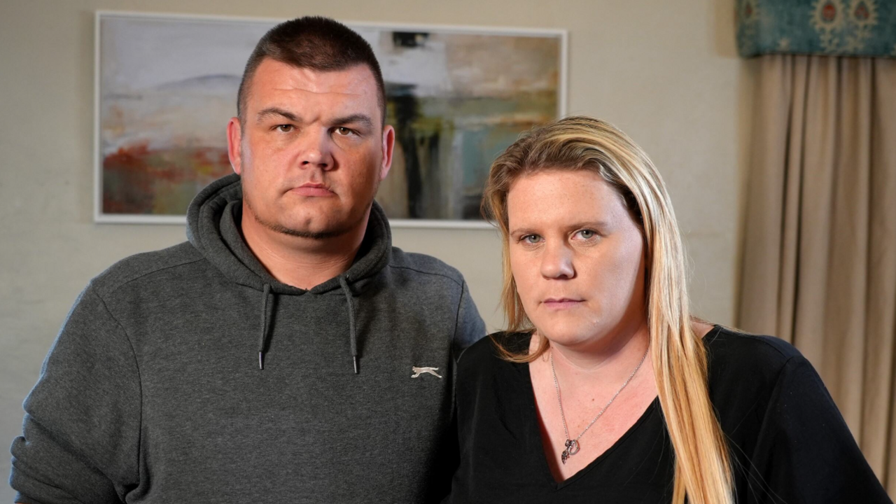 Lee and Micala Trussler stand side-by-side in a room with a painting on the wall behind them. Lee has short dark hair and is wearing a grey hoodie, Micala has long blonde hair and is in a black top.