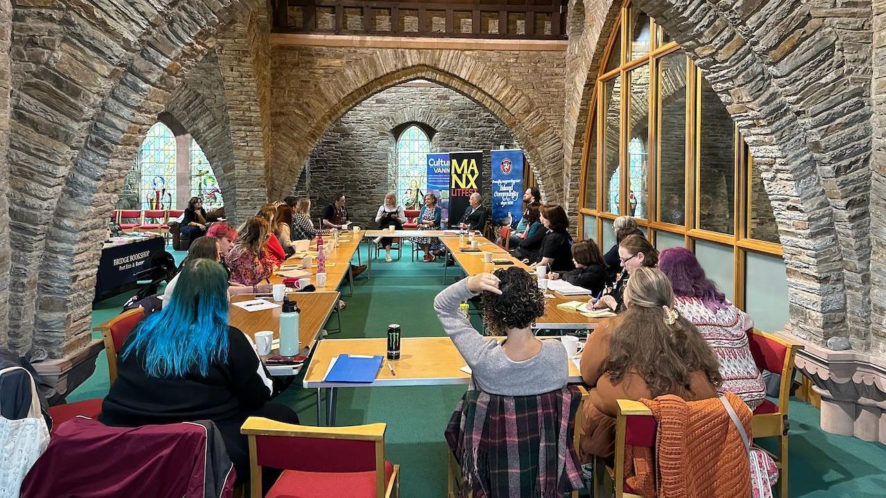 A long room with church style stone arches with tables grouped together in a circle and people sat round them