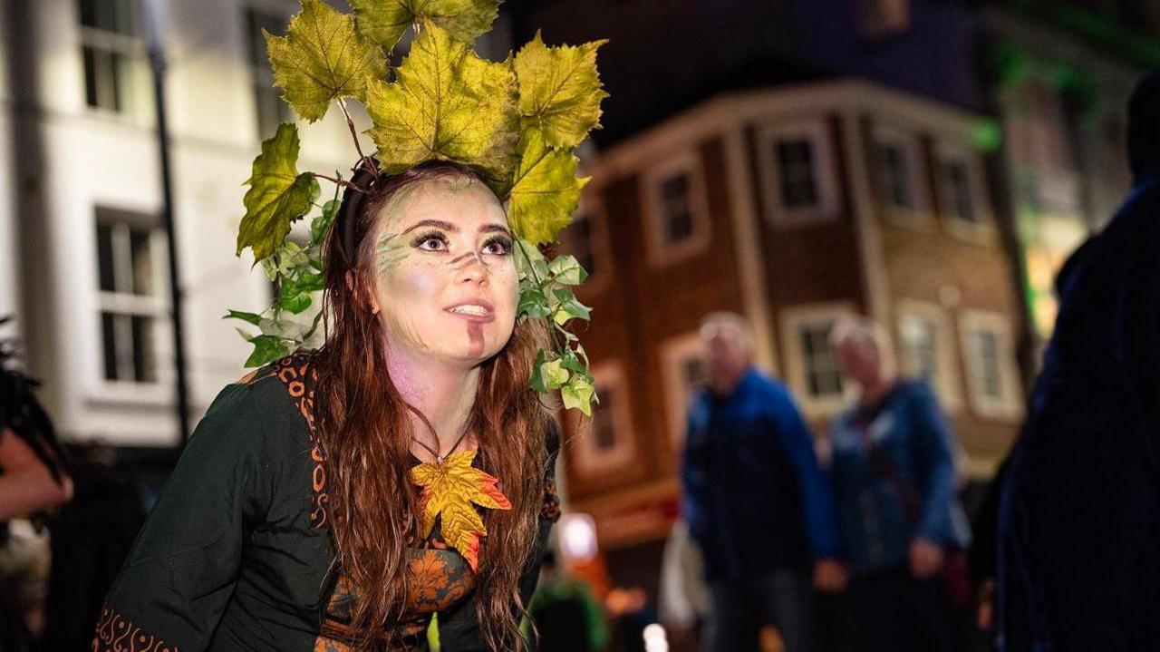 a woman in fancy dress, including a green dress and autumnal leaves, stands on a city centre street in Derry at Halloween