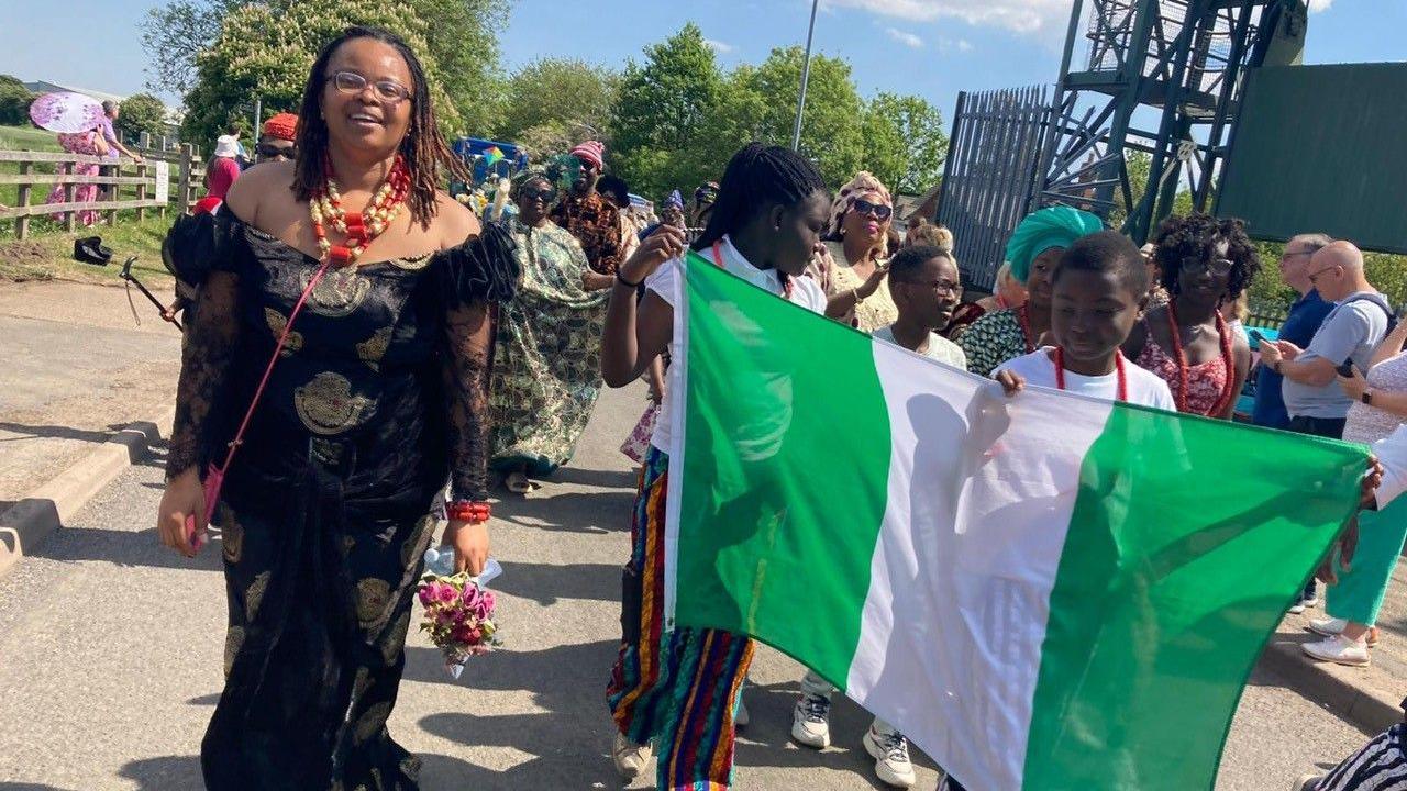Three youths carry a Nigerian flag to the right of the picture, and a lady wears a long black dress at the side and carries flowers and wears colourful beads round their neck.