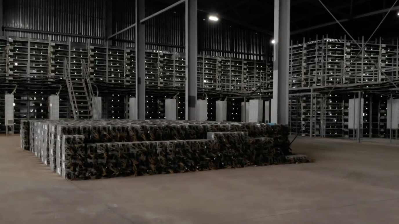 Black racks of powerful computers in a warehouse. They are surrounded by scaffolding and have white dotted lights on them.