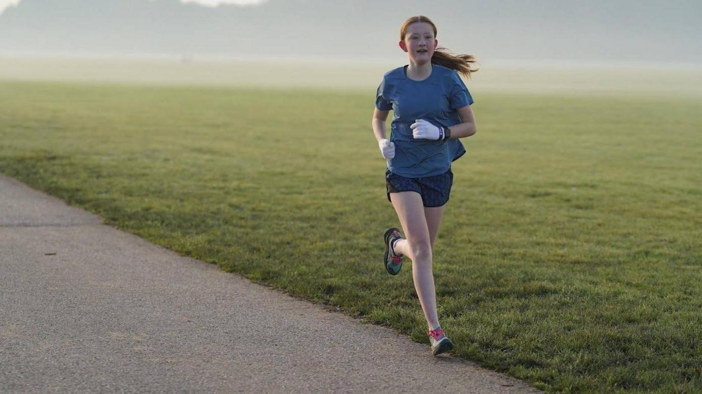 A teenage girl runs around a concrete track on York Knavesmire with a spread of grass beyond the track. She has long red hair tied up in a ponytail and wears a blue top, black shorts and light blue trainers with pink shoelaces.