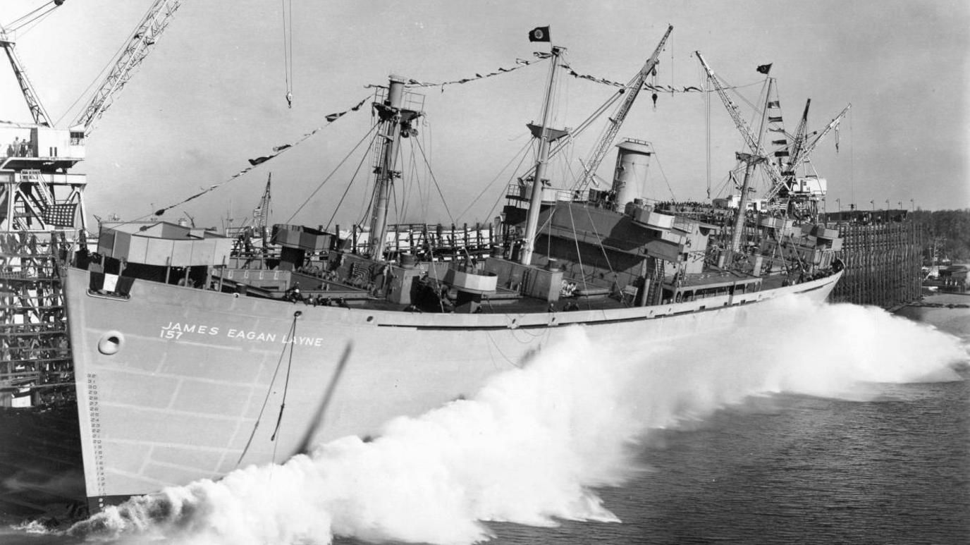 A black and white photograph shows the James Egan Layne Liberty Ship causing a wake as the boat is launched into the water for the first time in a dockyard 