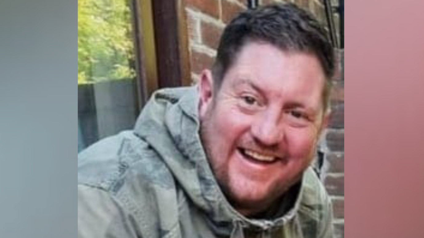 Colin Burns, wearing an camouflage jacket, sits while smiling at the camera. This photo is from several years ago and when he had an addiction to drugs and alcohol.