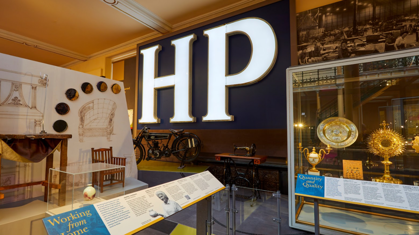 The letters HP are lit up on a wall with various other items in cabinets in the museum on display around it