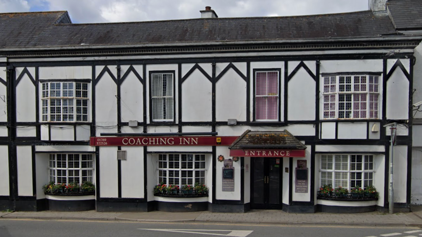 A Google Maps image capturing a white old fashioned building with black panelling, a door with ENTRANCE in red above it, to the left is COACHING INN sign with red background. Cloudy skies behind.