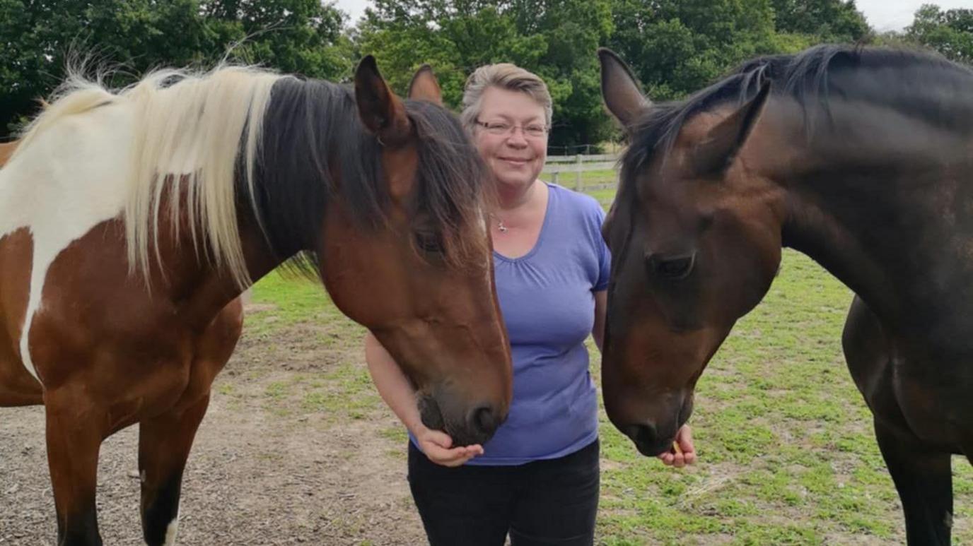 A woman wearing a lilac top while standing in a field with two horses. One is light brown with a brown and cream mane. The other is dark brown with a dark mane.