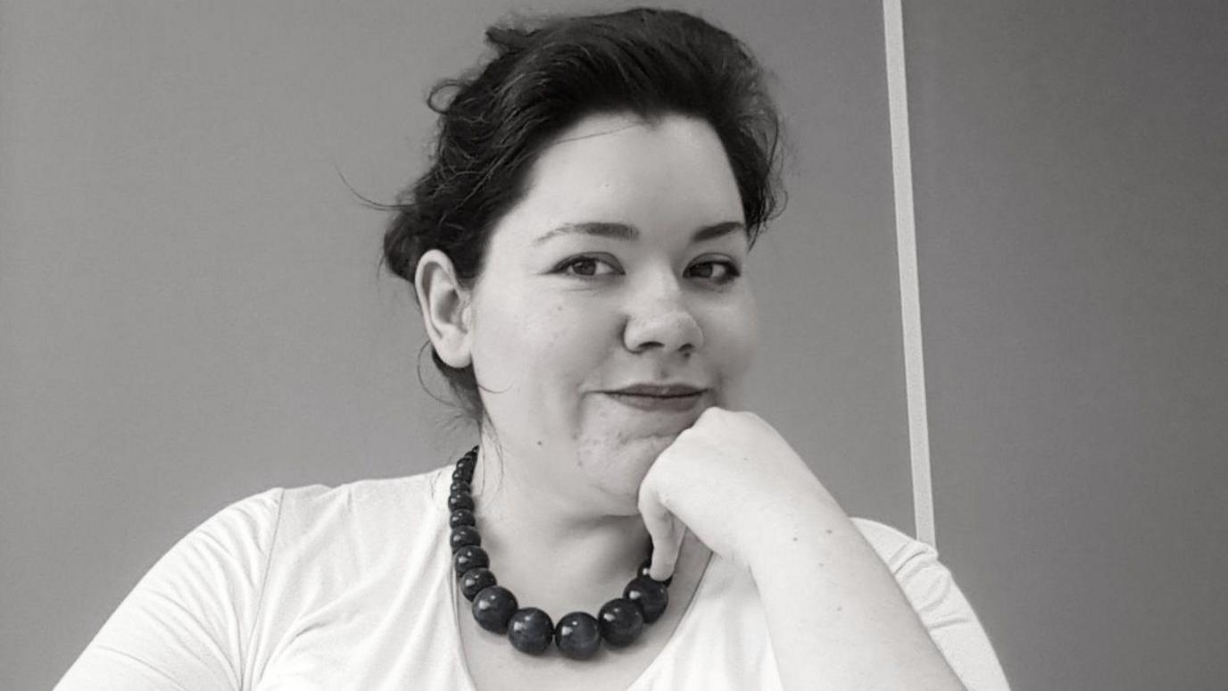 Rachael, who has dark hair, wearing a white t-shirt and a chunky bead necklace looks into the camera while resting her chin on her hand. The image is a black and white photo.