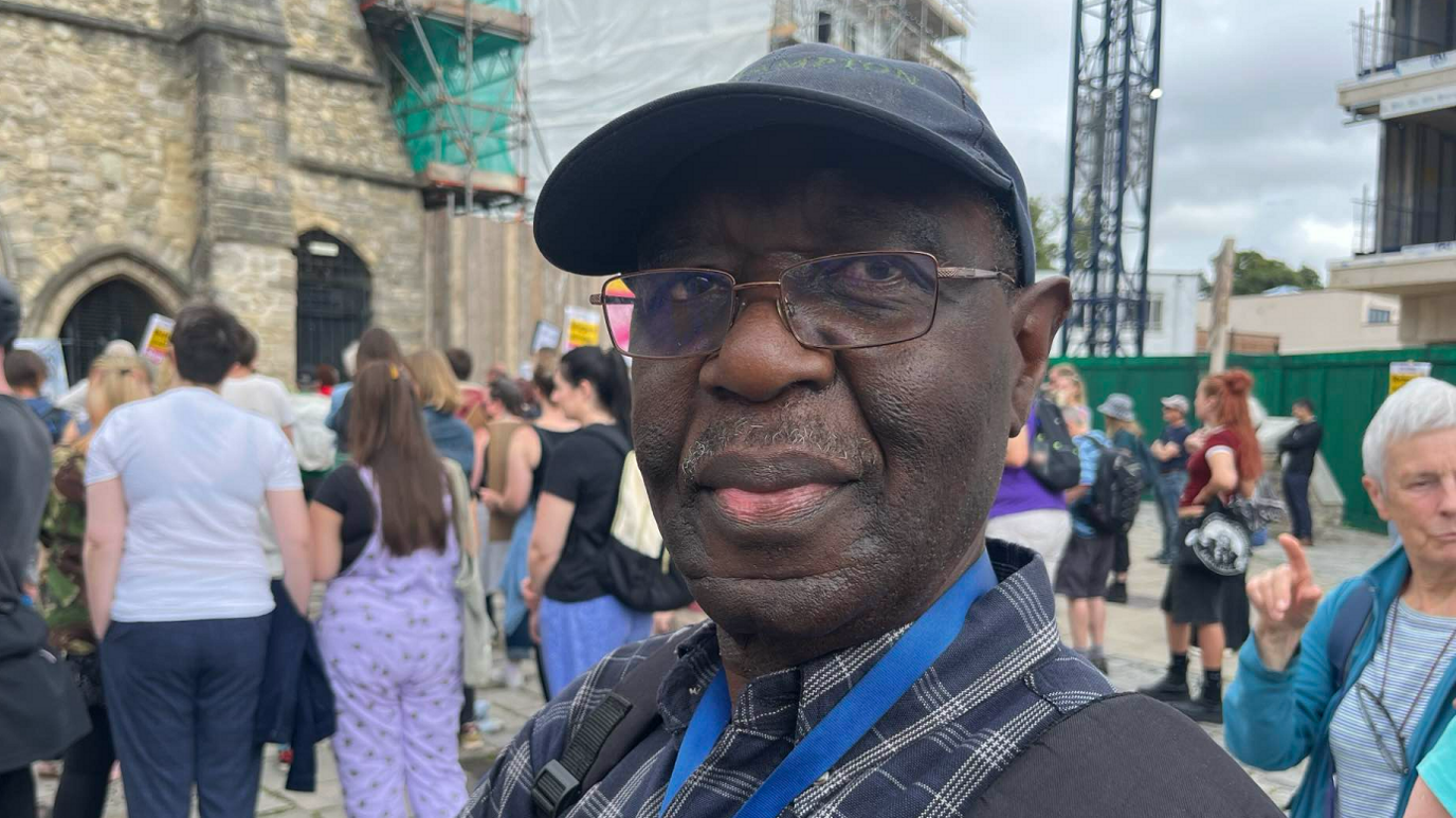 Prof Ademuni-Odeke is wearing a blue cap and a blue lanyard, a striped blue shirts and a black rucksack on his back