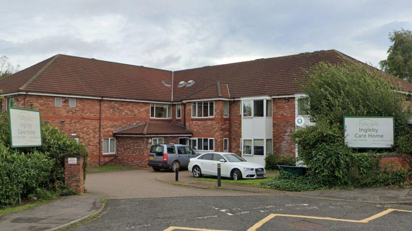 A general street view of Ingleby Care Home. The two-storey, L-shaped red brick building has a high slate roof, white windows and a single storey porch entrance in the corner of the "L". Tall hedges border the property and two cars are parked outside the building.