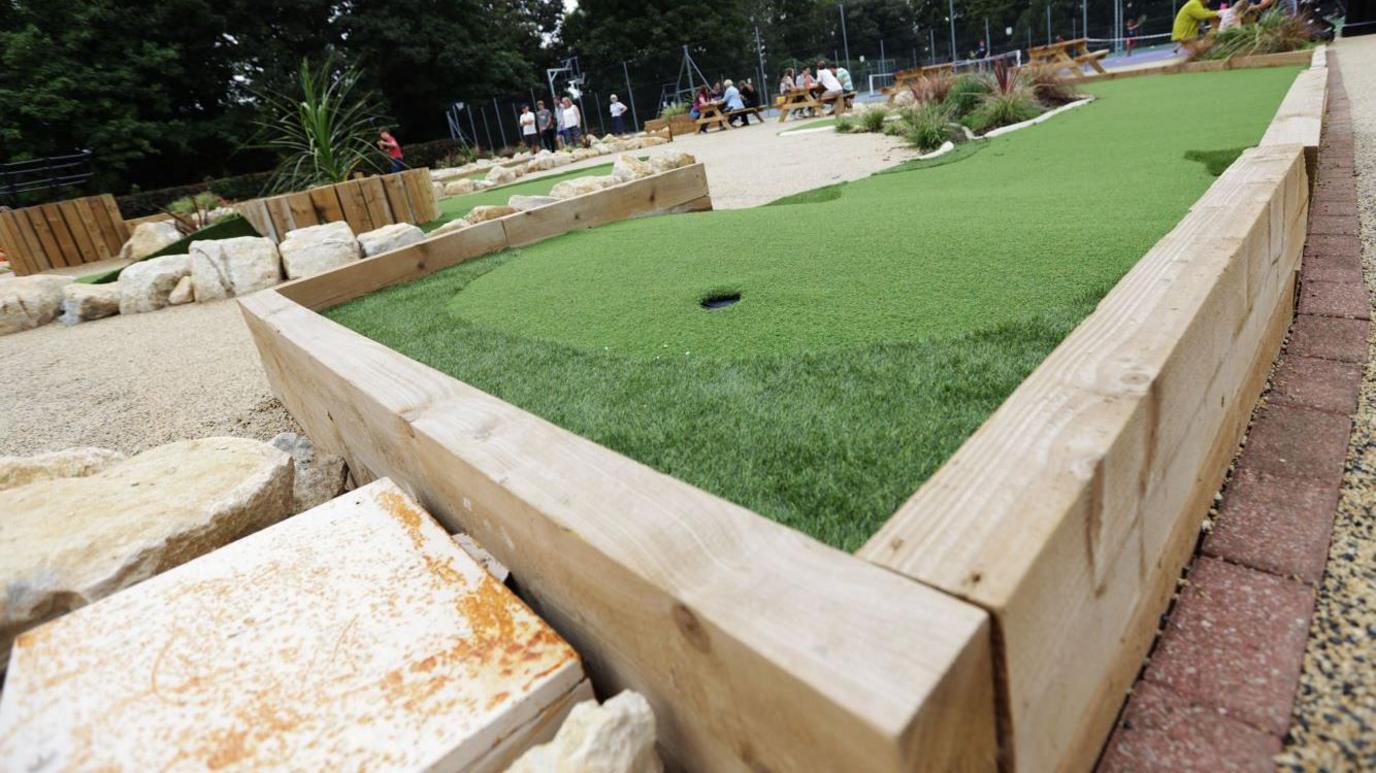 A mini golf course, one of the holes is close to the camera. People can be seen sitting at tables in the distance. It's an overcast day.