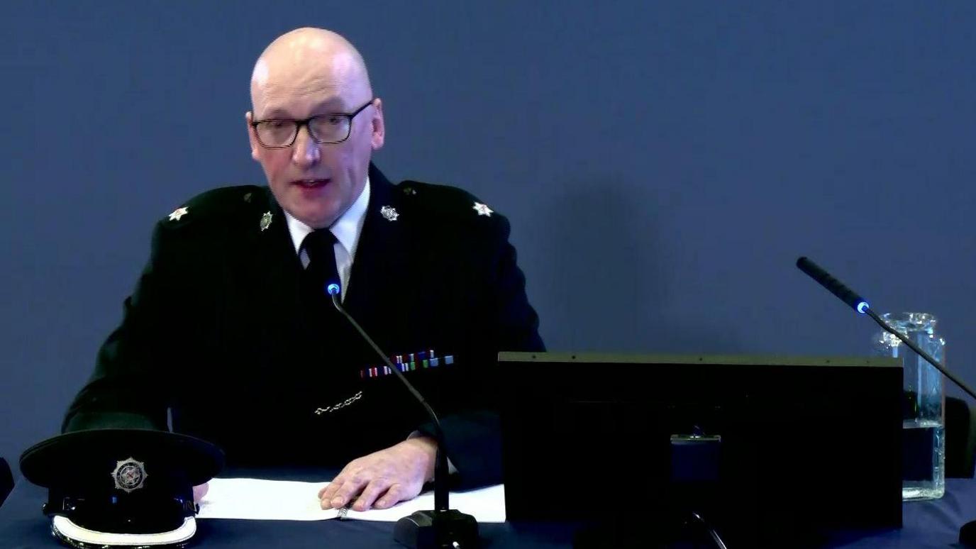 Norman Haslett at the Omagh Bombing Inquiry. He is wearing a police uniform and sitting in front of a computer and microphones. He is bald and wears glasses.