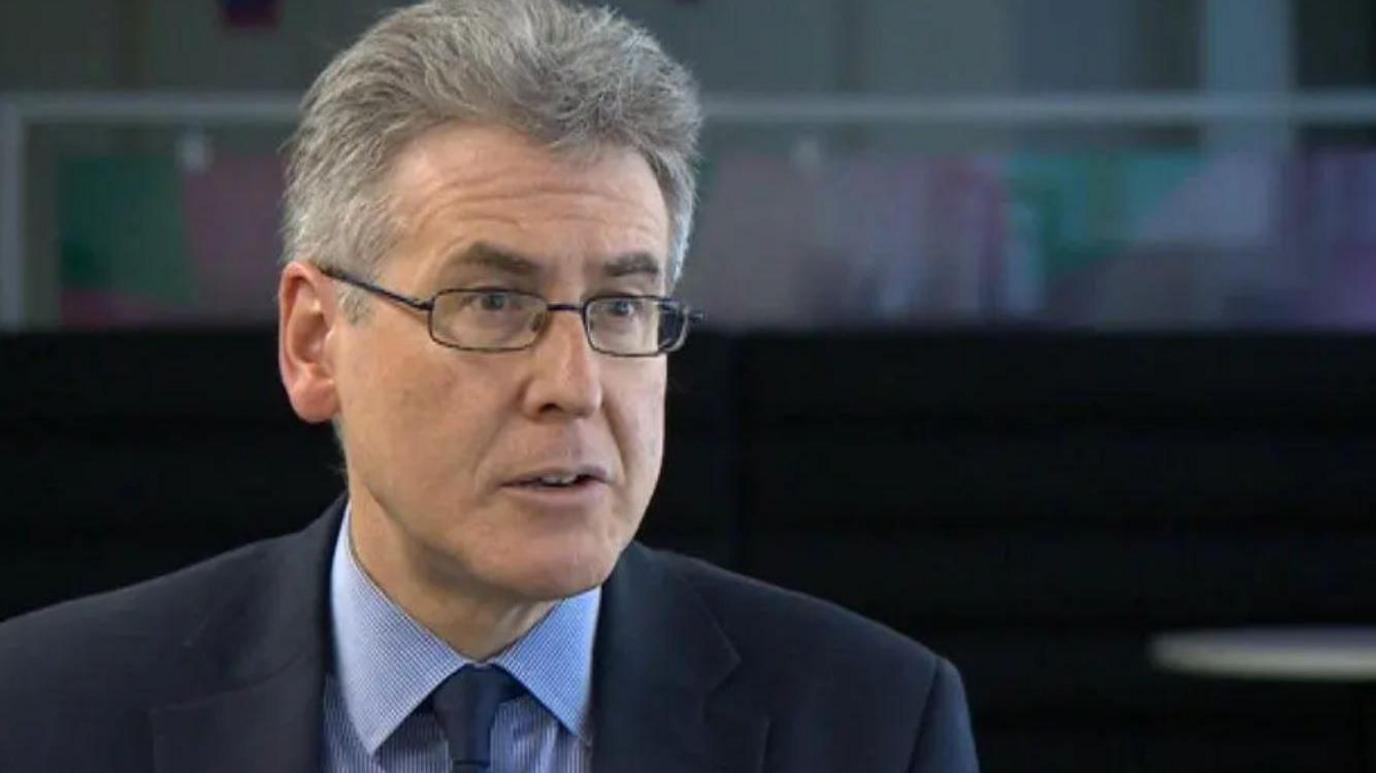 A man with grey hair and glasses in a dark suit and tie