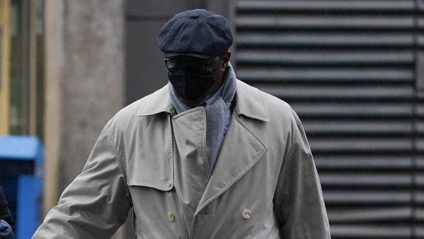 Charles Ehikioya wearing a beige trench coat, grey scarf, glasses, a black mask covering his face and a grey hat.