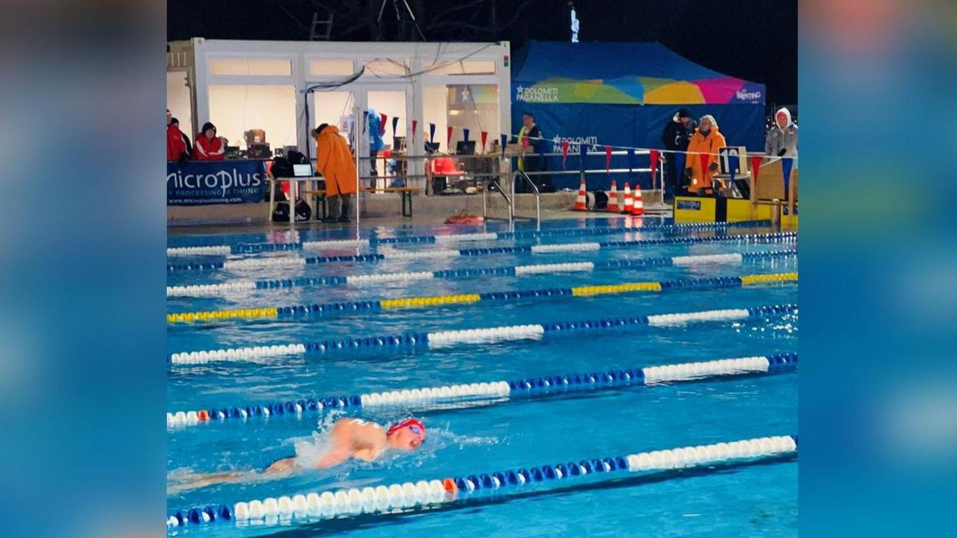 Helen Hislop is swimming while a number of people at the side of the pool are wrapped up against the freezing weather, wearing coats and hats.