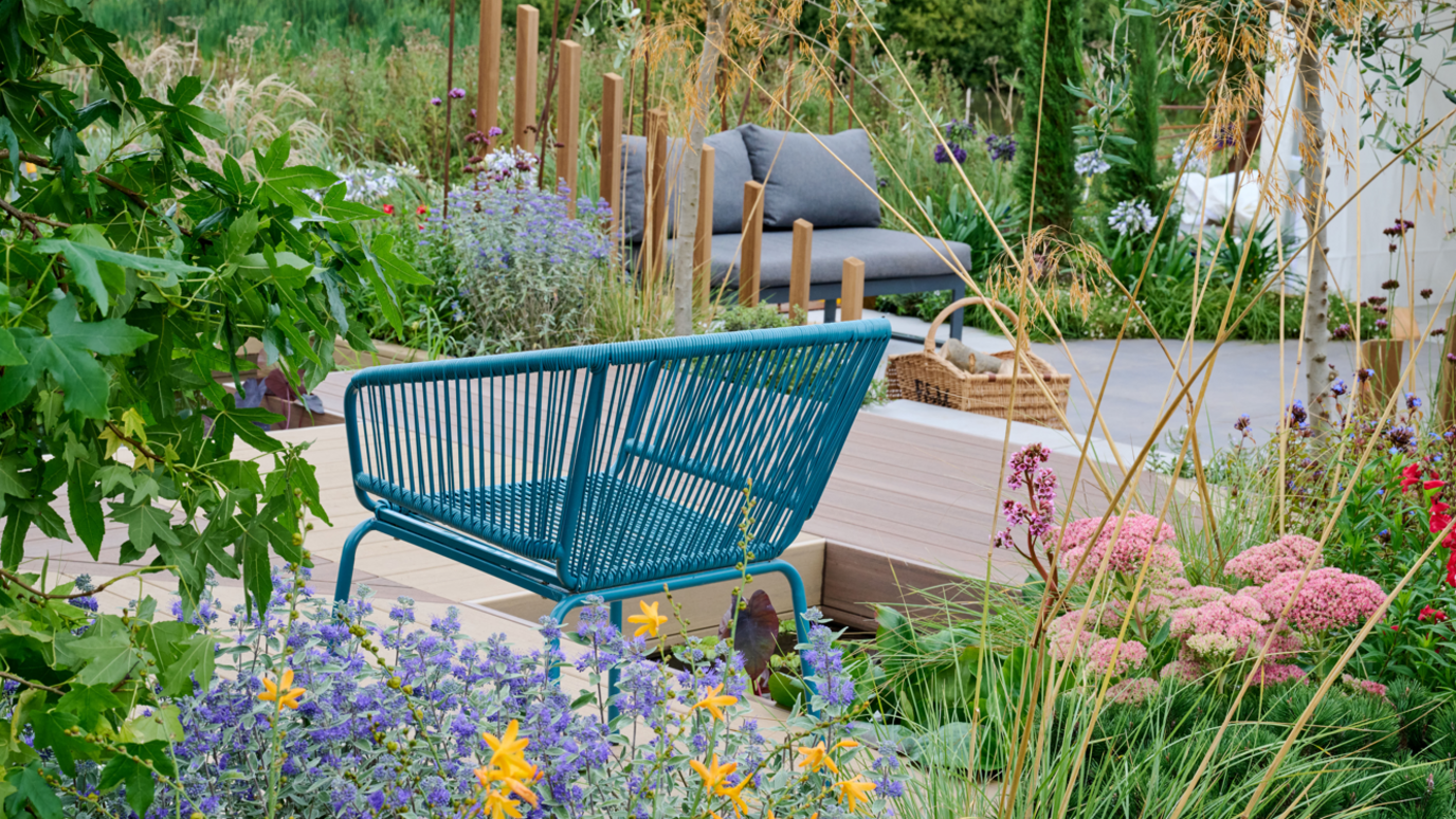 A blue wicker chair is to the left of the image on light brown wooden decking. In the background is a grey seat which has two cushions. There are different coloured plants planted around the seating areas.