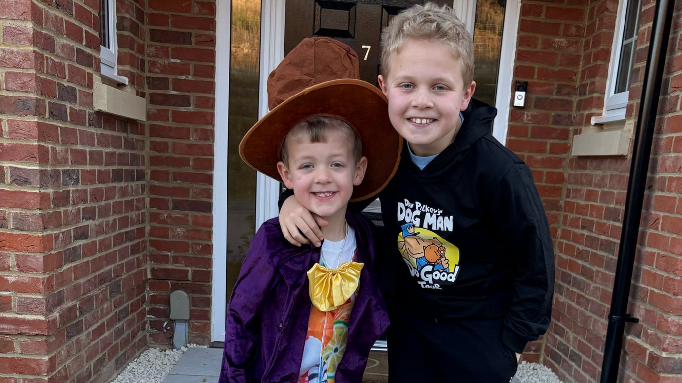 Charlie is wearing a purple jacket and a bow tie and a big brown hat and George is stood next to him, with his arm around Charlie and is wearing a black Dog Man hoodie. 