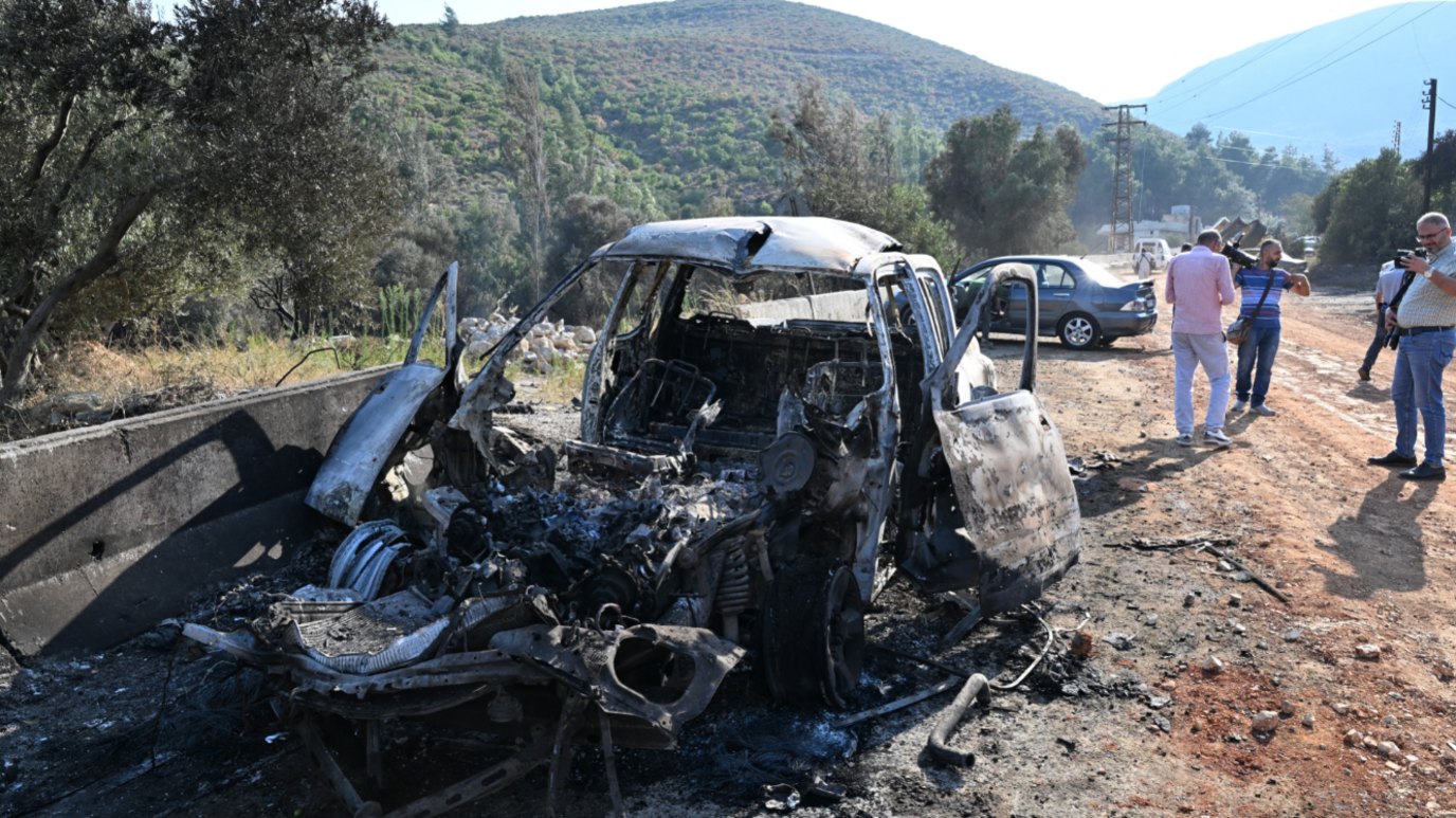 A damaged burnt out car on the outskirts of Masyaf lies on the side of a dirty road in Syria. People watch on and take photos of the damaged car.