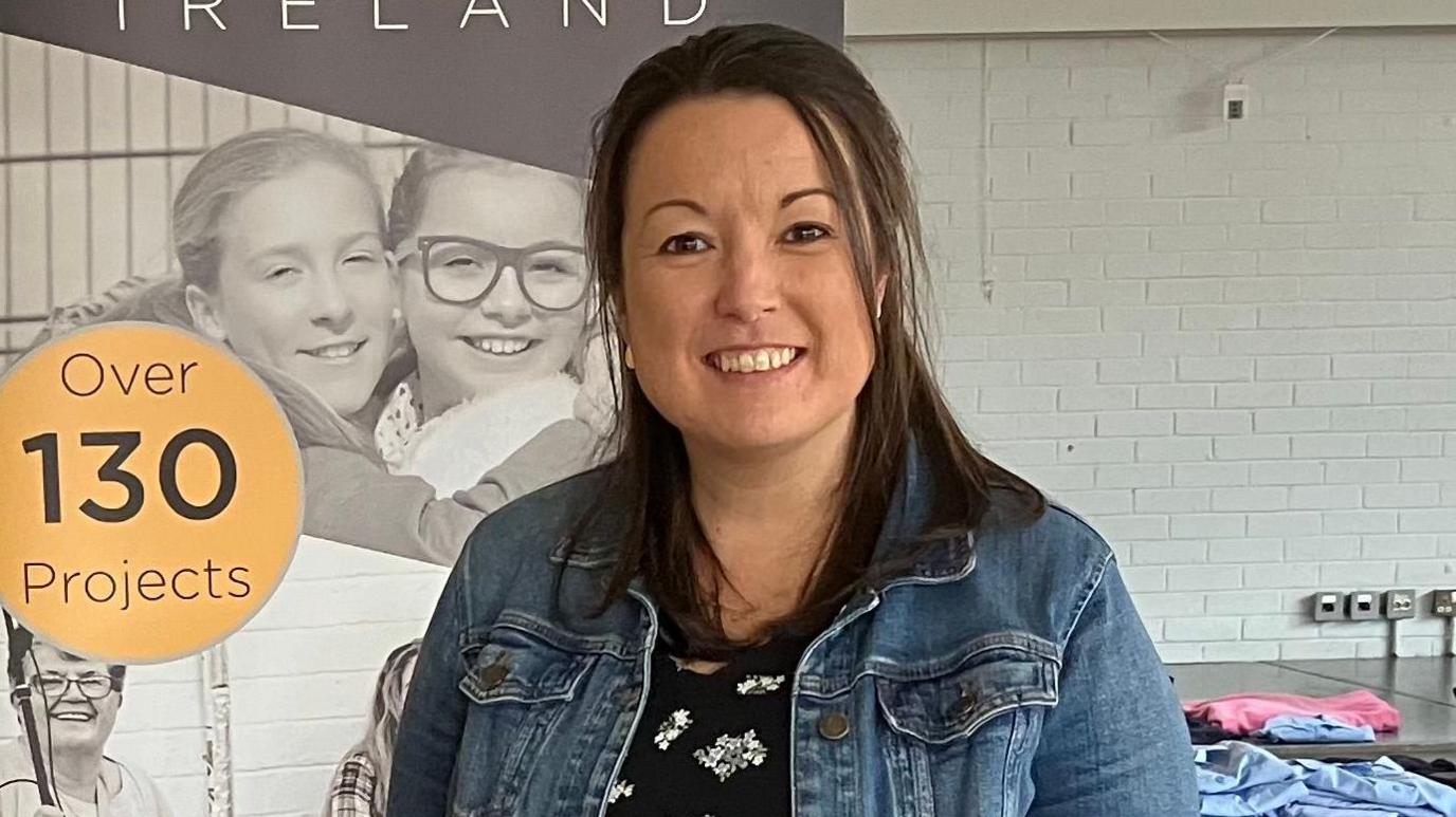 A woman with medium length dark hair smiles at the camera. She is wearing a denim jacket and a black floral top and in the background there are piles of school uniform polo shirts.