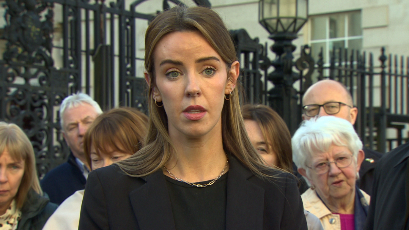 Marian Vincent has long brown hair and blue eyes and is wearing a dark suit jacket and top. She is speaking with other relatives of the dead standing behind her