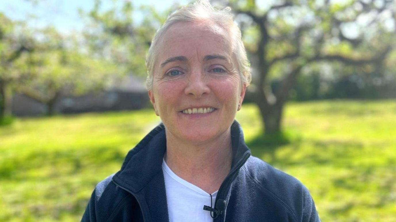 Rose has fair hair pulled back. She's smiling into the camera and is wearing a navy hoodie with a white top. She's standing on grass and there are trees behind her.