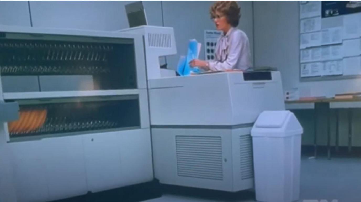 A still image from the film 9 to 5. Jane Fonda's character stands next to a huge Xerox photocopying machine, which is bigger than she is. The grey L-shaped machine has one open side with slots for paper and a closed side with grills. Jane has short curly coiffed hair and wears large glasses and a lilac blouse. She holds a pile of papers and is looking anxiously at the machine.