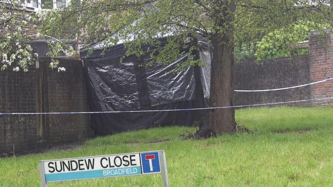 A black forensics tent surrounded by blue and white police tape stands beyond a tree on a green in a residential area 