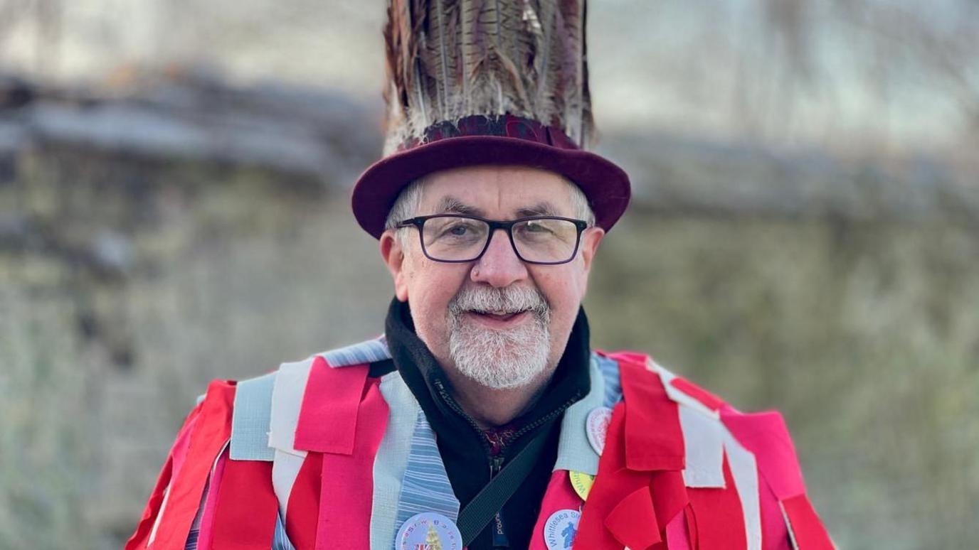 Gary Cook in a red shawl. He is looking at the camera and is wearing black rimmed glasses and a hat with feathers. 