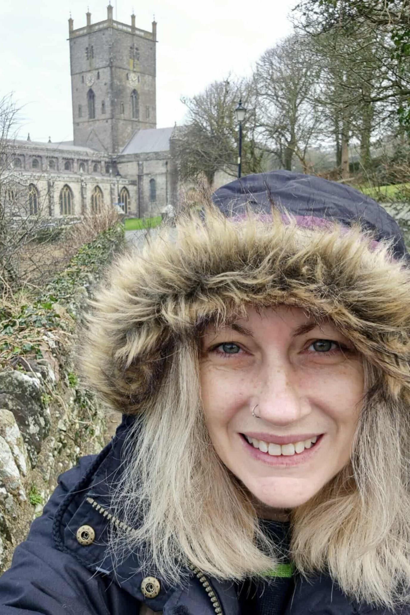 A woman with blonde hair with a black coat on with a faux-fur hood up. She smiles at the camera as she takes a selfie. Behind her is a Cathedral. 