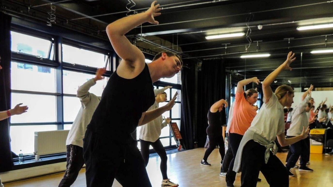 A group of people in a dance studio. They have their arms above their head and are leaning over to one side.