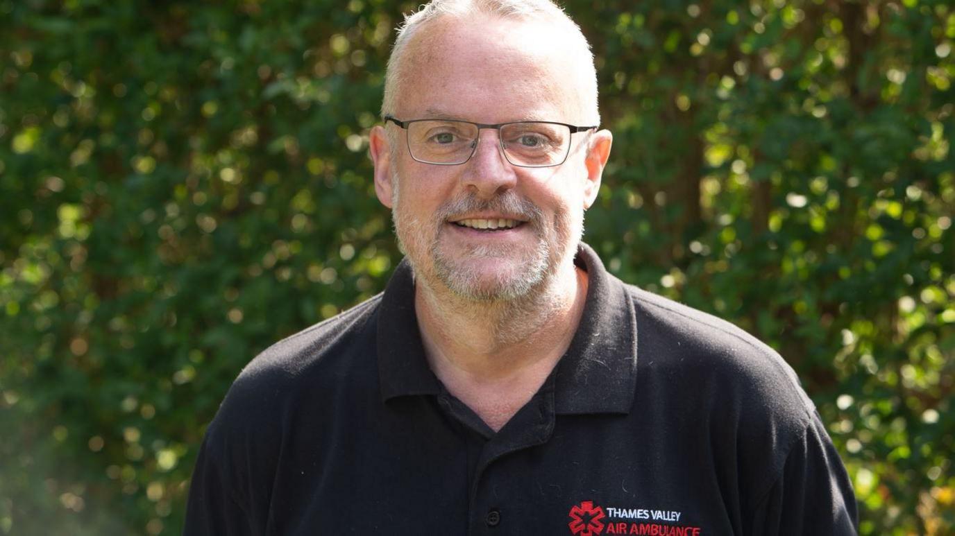 Adam Crosby smiling for the camera outside on a sunny day. He is wearing a black top branded with the Thames Valley Air Ambulance logo. There is greenery behind him. 