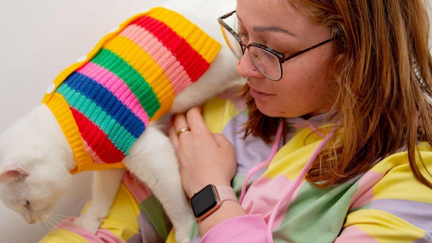 A woman in a striped hoodie with a white cat walking from her shoulder down her arm, also wearing a rainbow striped pullover. The woman has red hair and is wearing glasses and a nose stud.