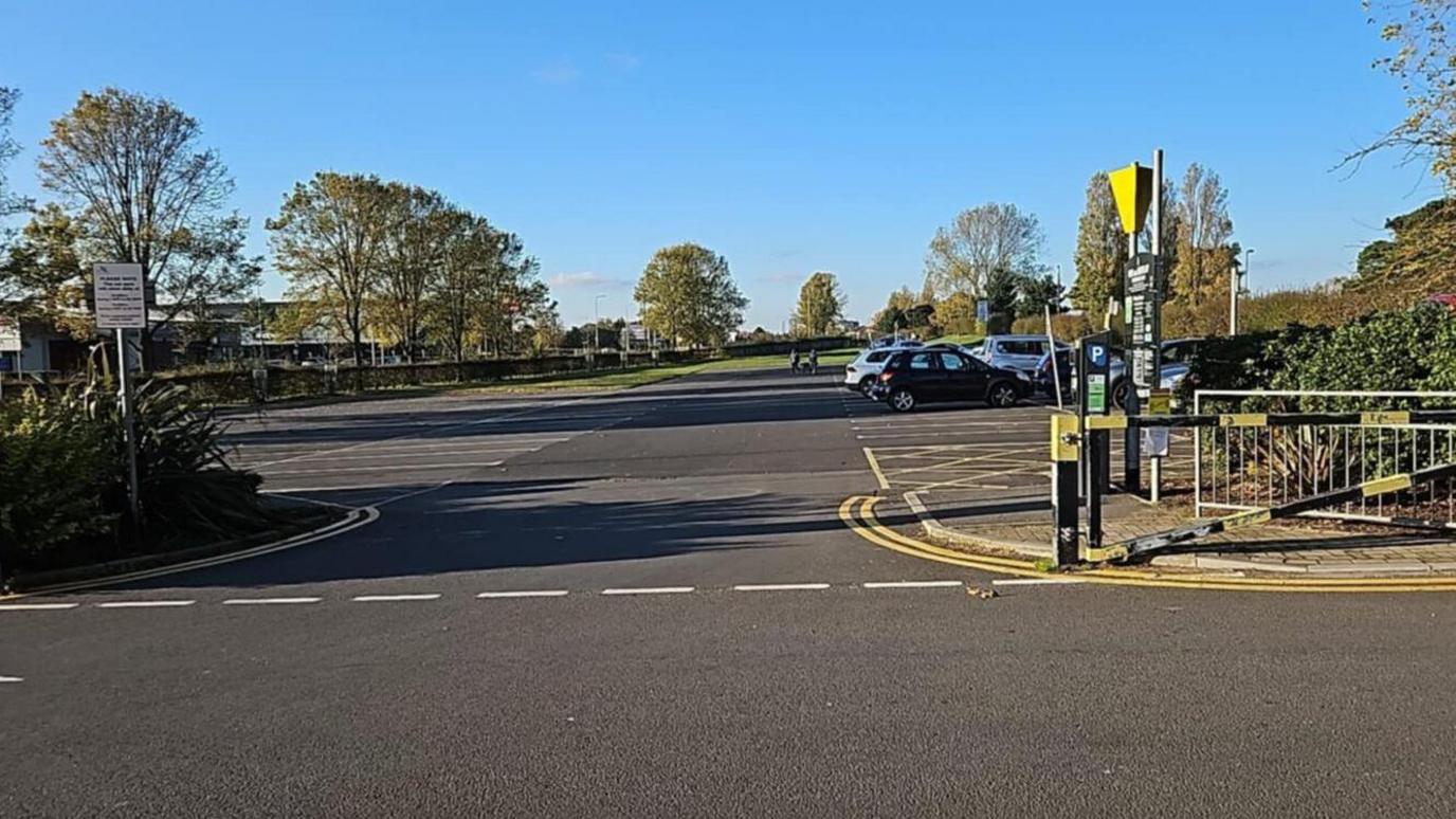 The entrance to the car park at Cleethorpes' boating lake. It is a large space with a pay and display machine. There are trees and fields in the distance.