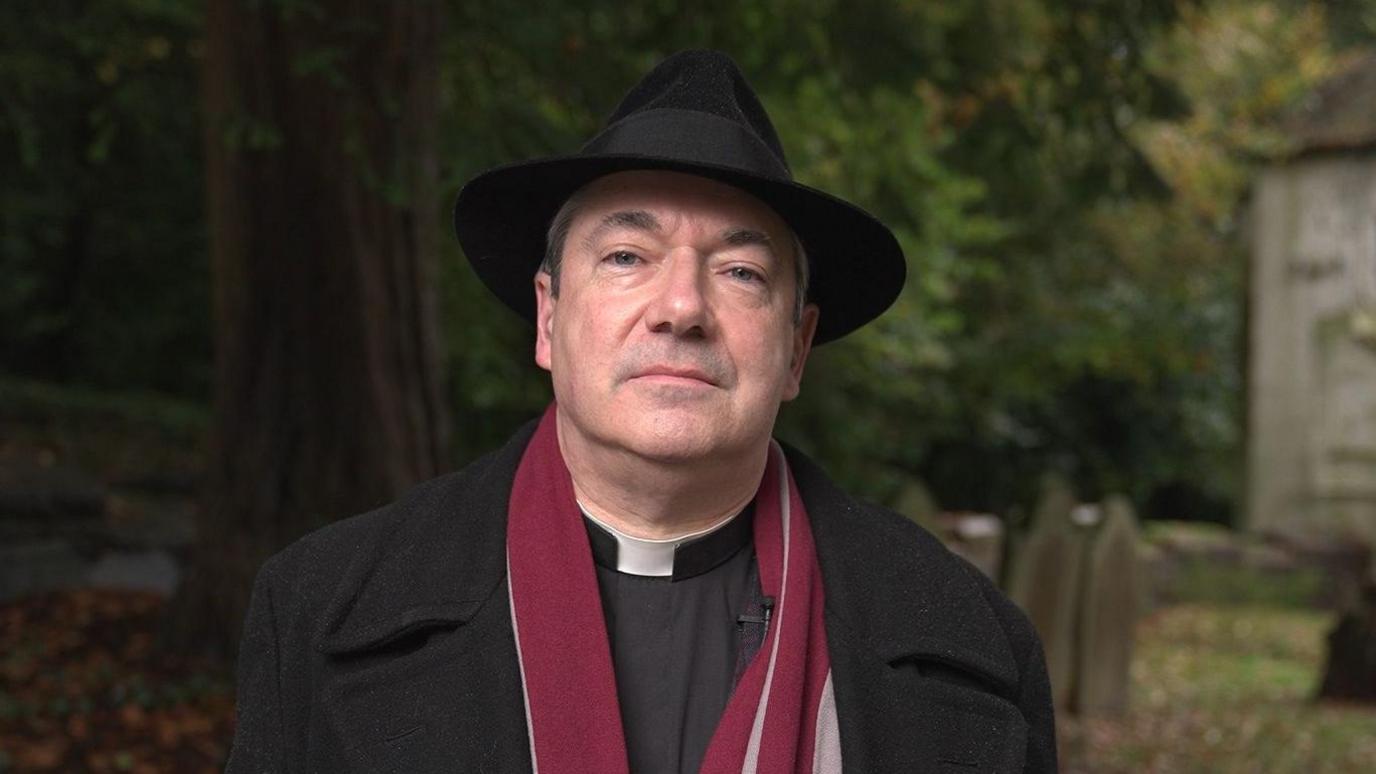 A man wearing a trilby, long black coat, and burgundy scarf stands beneath a tree, in front of several aged headstones.