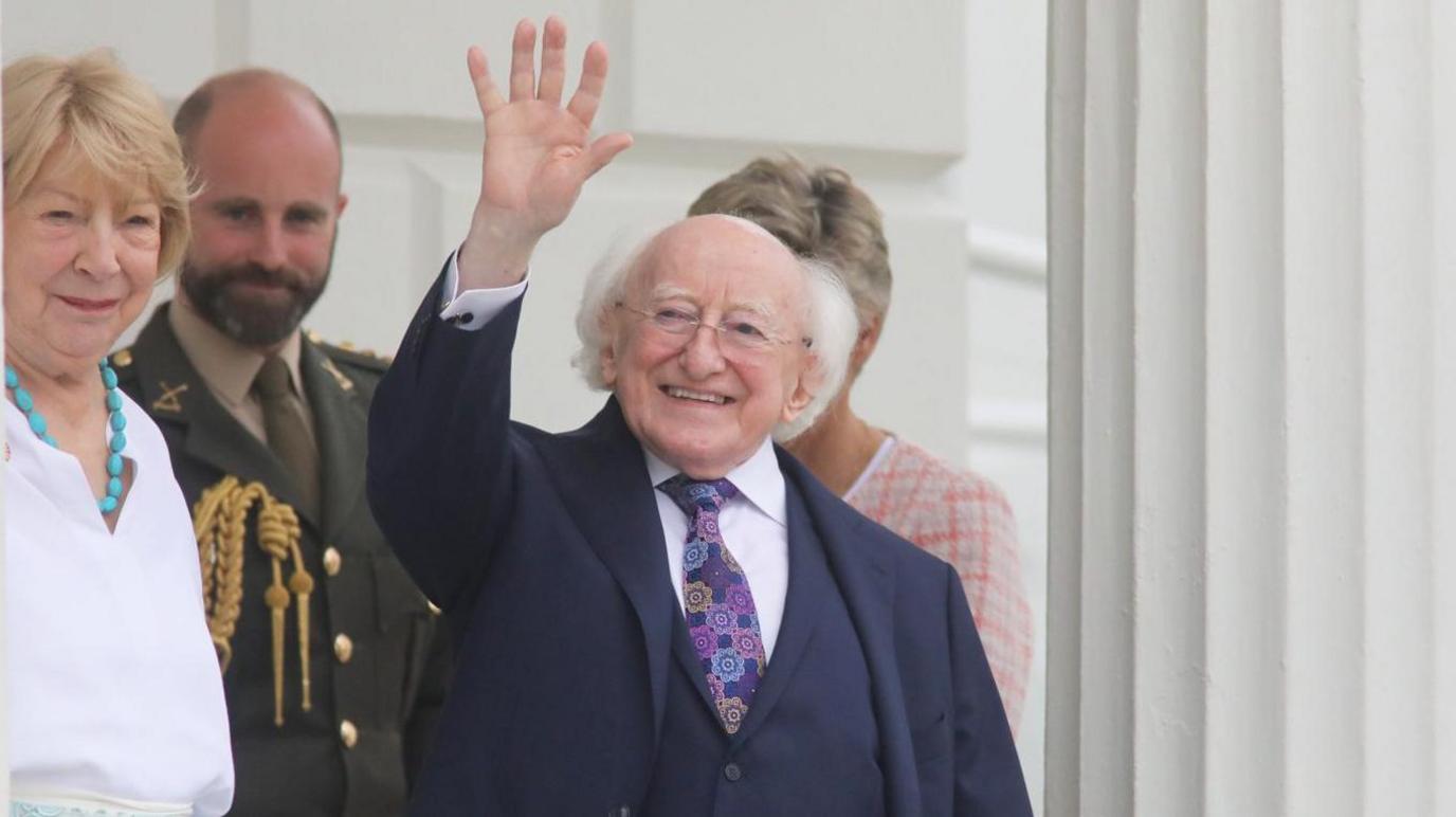 Michael D Higgins smiles as he waves at the camera. He is bald on the top of his head and has grey hair at the side. He is wearing a navy suit with a white shirt and multicoloured tie. His wife and other people are standing behind him.